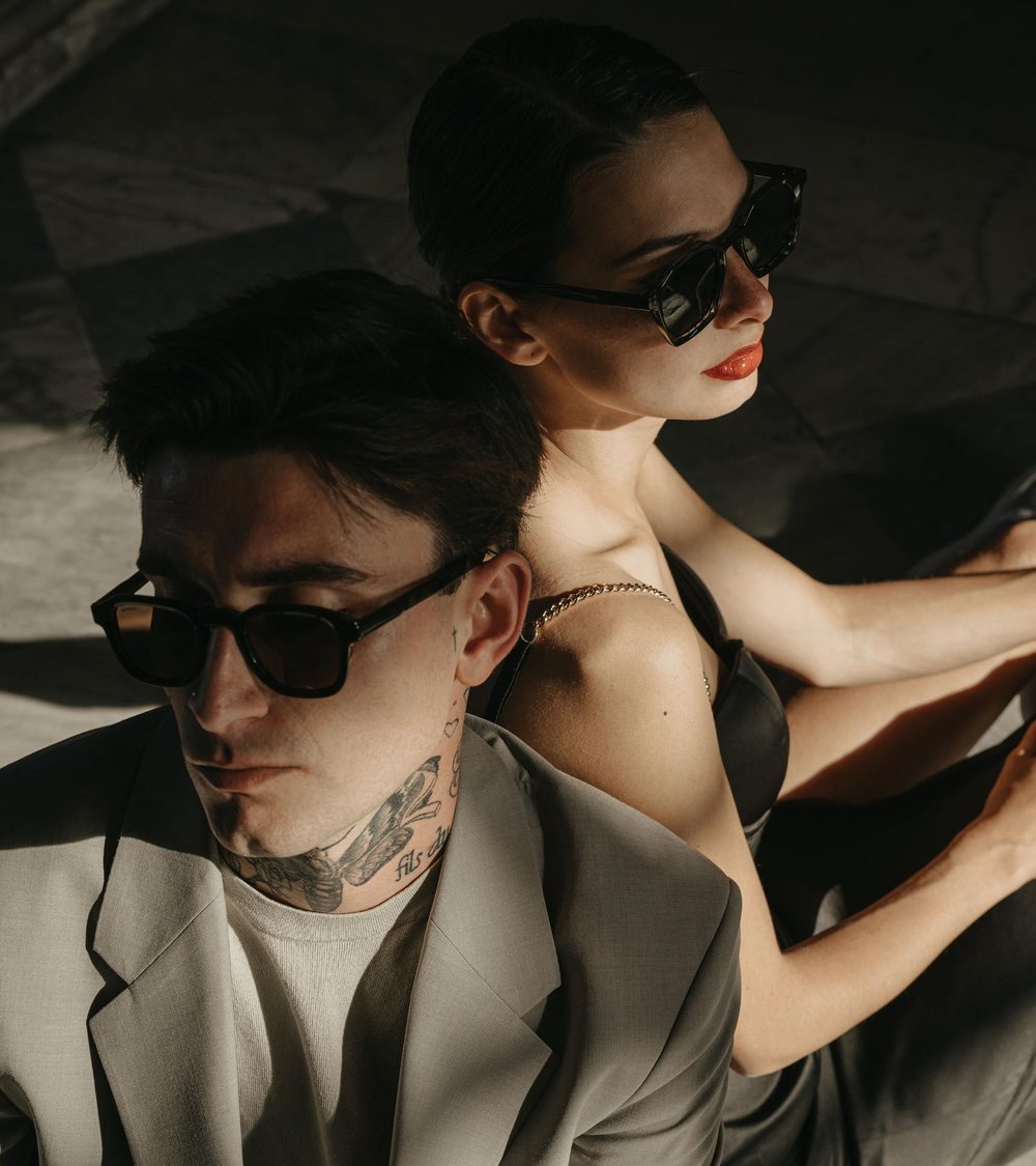 A man and a woman seated back to back, both wearing Moncada sunglasses